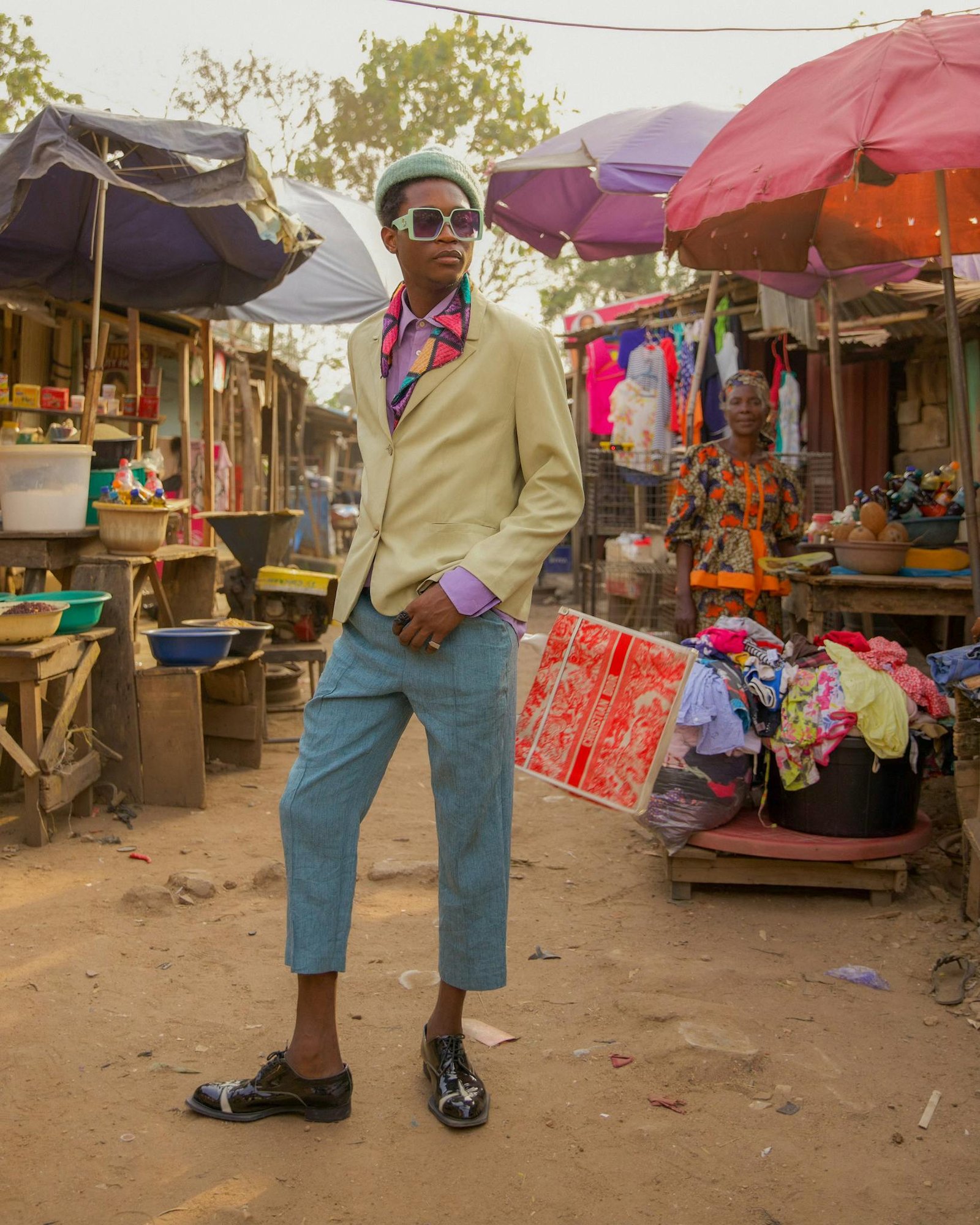 A fashionable man in colorful attire stands confidently in a bustling outdoor market.